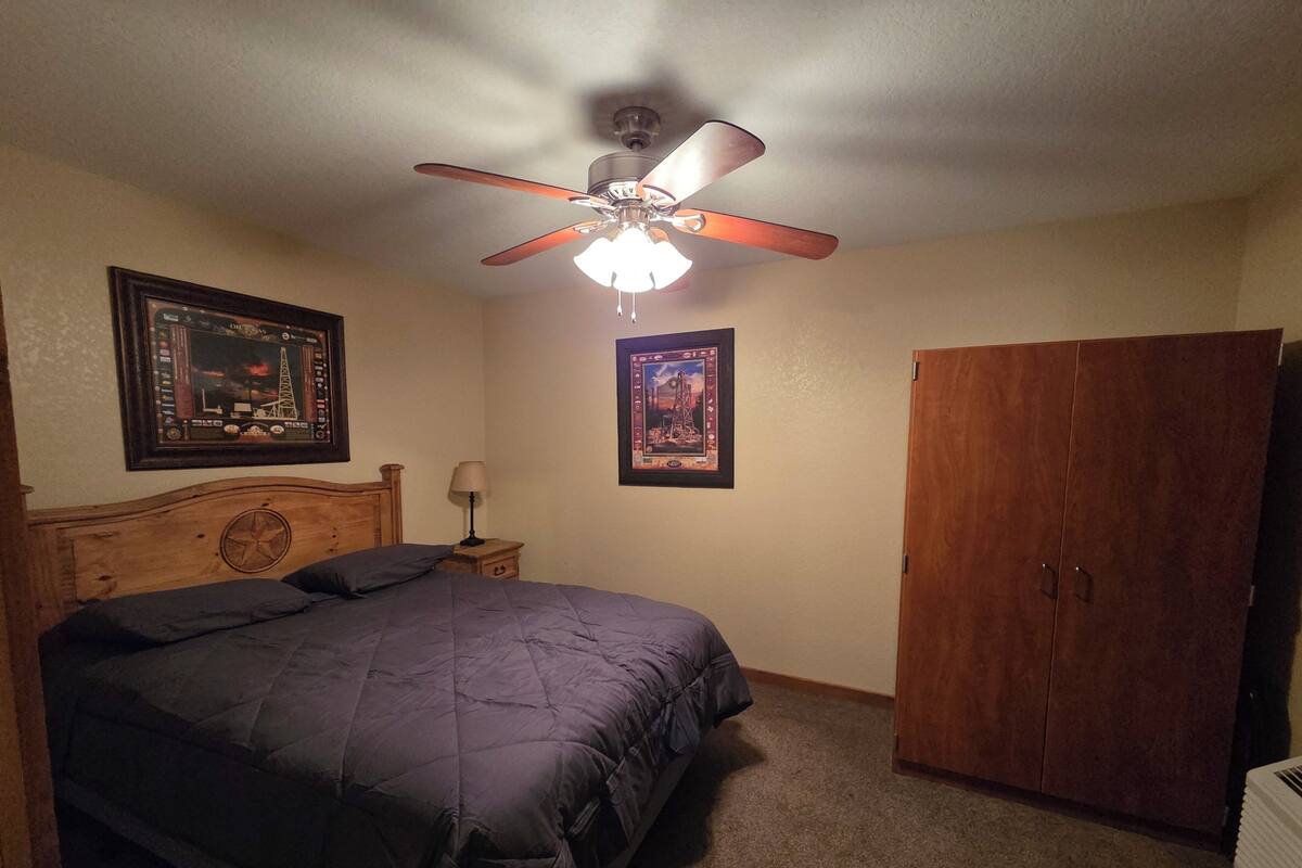 A bedroom with a bed, a wooden wardrobe, a ceiling fan, and two framed pictures on the wall.