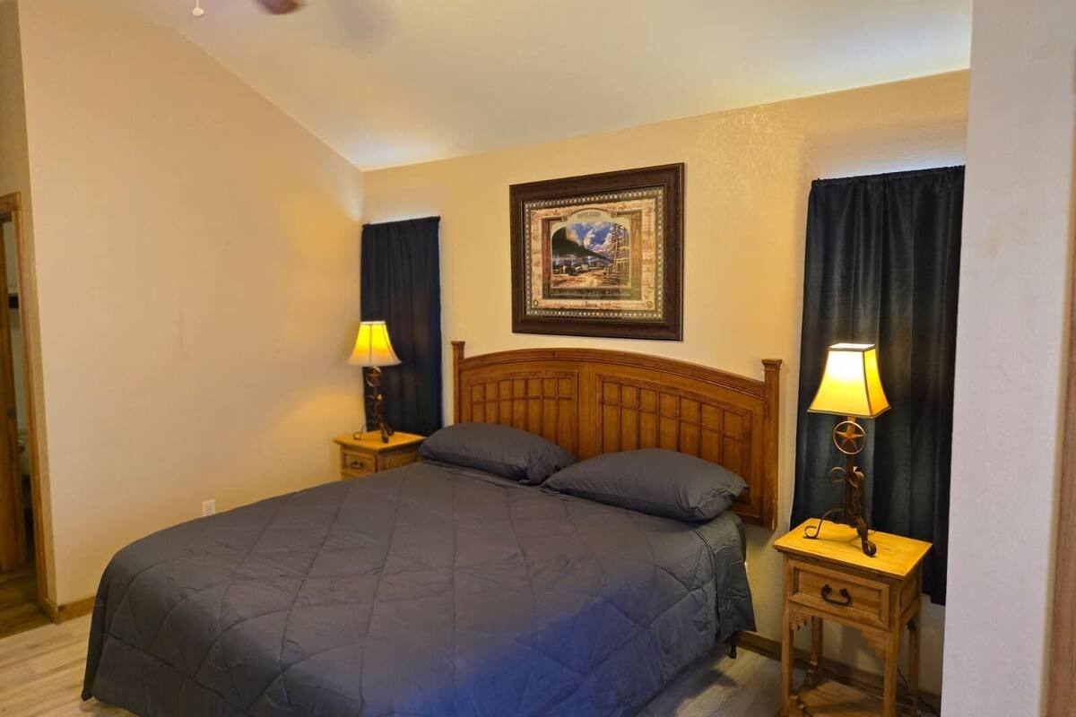 A neatly made bed with a quilted blue comforter and pillows, flanked by two bedside tables with lamps, a framed picture on the wall, and a ceiling fan above.