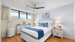 Ein Schlafzimmer mit einem Bett, einem Deckenventilator, einem Nachttisch mit Lampe, einem Schminktisch und einem Fenster mit Vorhängen.