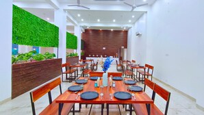 A modern dining area with wooden tables and chairs, green wall decorations, and ceiling fans. - Foreste Resort (Ramnagar)