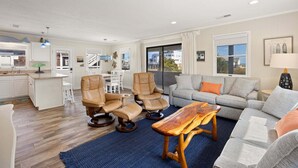 A spacious living room with a grey sofa, two leather chairs, a wooden coffee table, and a kitchen area with white cabinets and a dining table. - Little Island Time: Walk to Beach Retreat (Kitty Hawk)