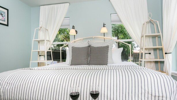 A bedroom with a large bed, striped bedding, and two bedside tables with lamps. - Dogologie Haus | 2/2, Pet Amenities, Gas Grill, Pets w/ Fee (Fredericksburg)