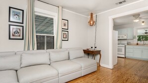 A modern living room with a light-colored sectional sofa, a wooden floor, and a kitchen area with white cabinets and appliances. - Dogologie Haus | 2/2, Pet Amenities, Gas Grill, Pets w/ Fee (Fredericksburg)