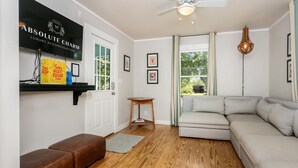 A living room with a beige sofa, a brown leather ottoman, a wooden coffee table, and a ceiling fan. There is a black shelf with books and a framed sign on the wall. - Dogologie Haus | 2/2, Pet Amenities, Gas Grill, Pets w/ Fee (Fredericksburg)