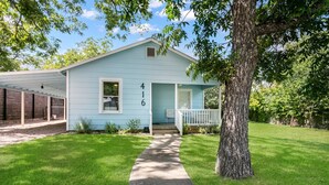 A light blue house with a number '416' on the front, a porch, and a tree in front. - Dogologie Haus | 2/2, Pet Amenities, Gas Grill, Pets w/ Fee (Fredericksburg)