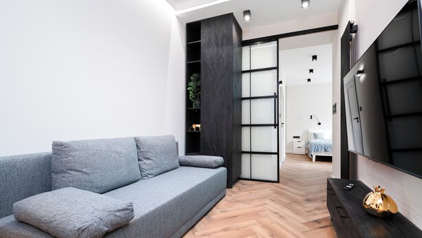 A modern living room with a grey sofa, a wooden herringbone floor, a black TV stand, and a sliding door leading to a bedroom. - Piotrkowska Grand Avenue Apartment #C (Łódź)