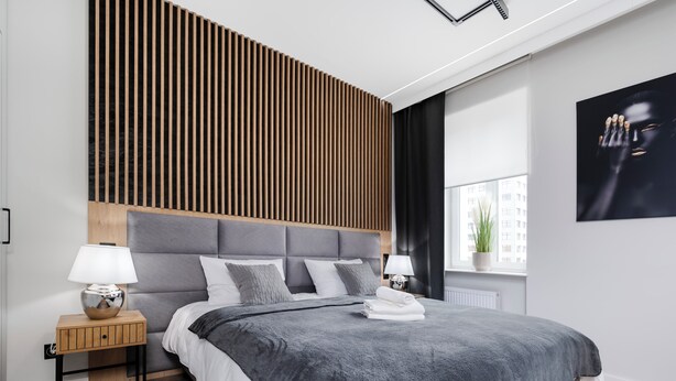 A modern bedroom with a grey upholstered headboard, a grey fluffy bedspread, and a wooden nightstand with a lamp.