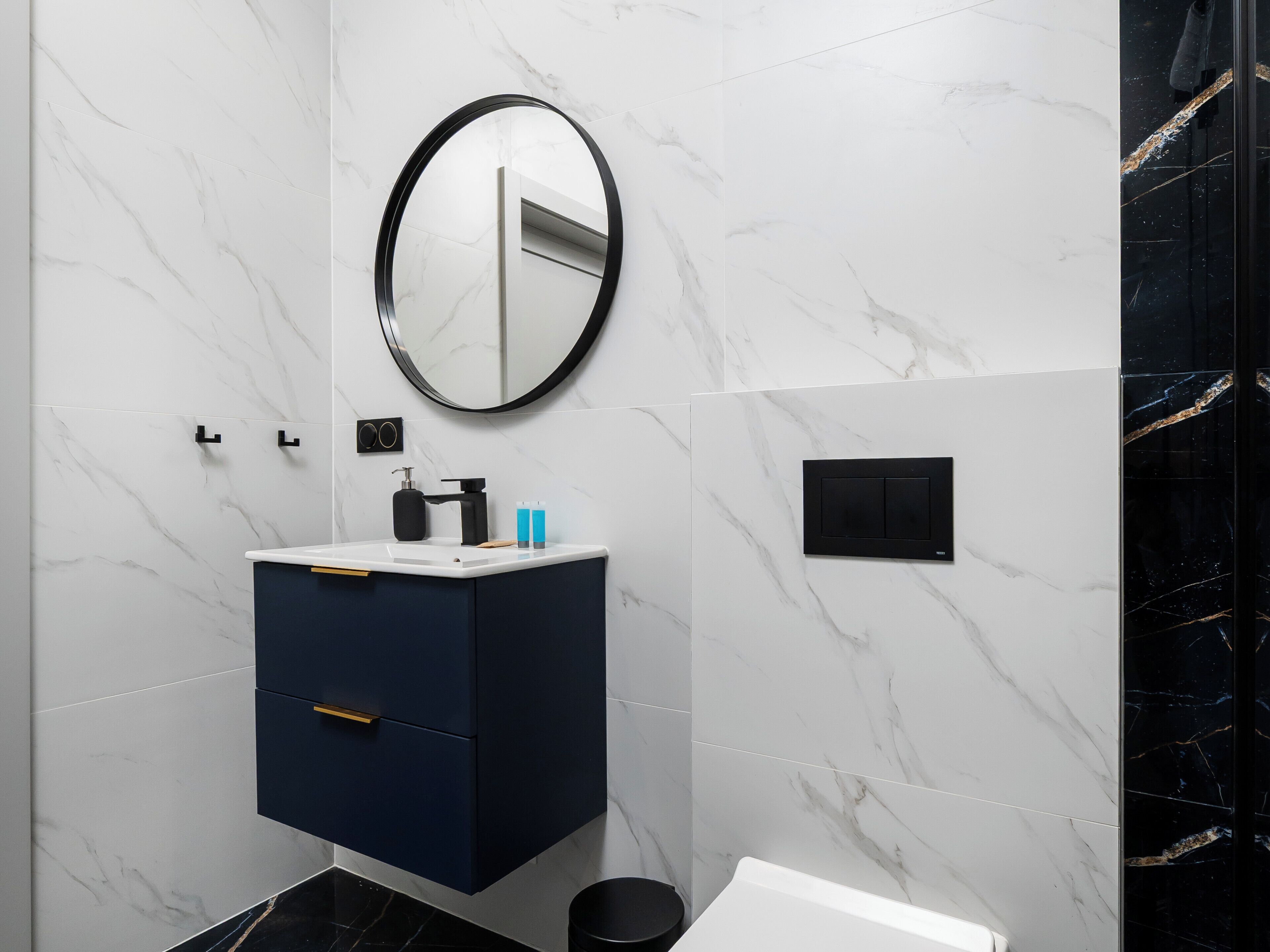 A modern bathroom with a marble wall, a round mirror, a dark blue cabinet, and a black sink.