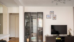 A modern living room with a wooden floor, a glass-fronted cabinet with decorative items, a flat-screen TV, and framed pictures on the wall. - Warszawa Wola Homely Apartment (Warszawa)