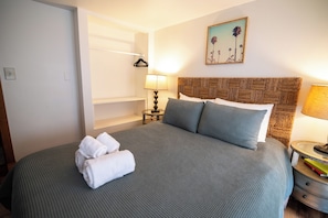 A neatly made bed with a grey bedspread and pillows, a bedside table with a lamp, and a wall-mounted picture.