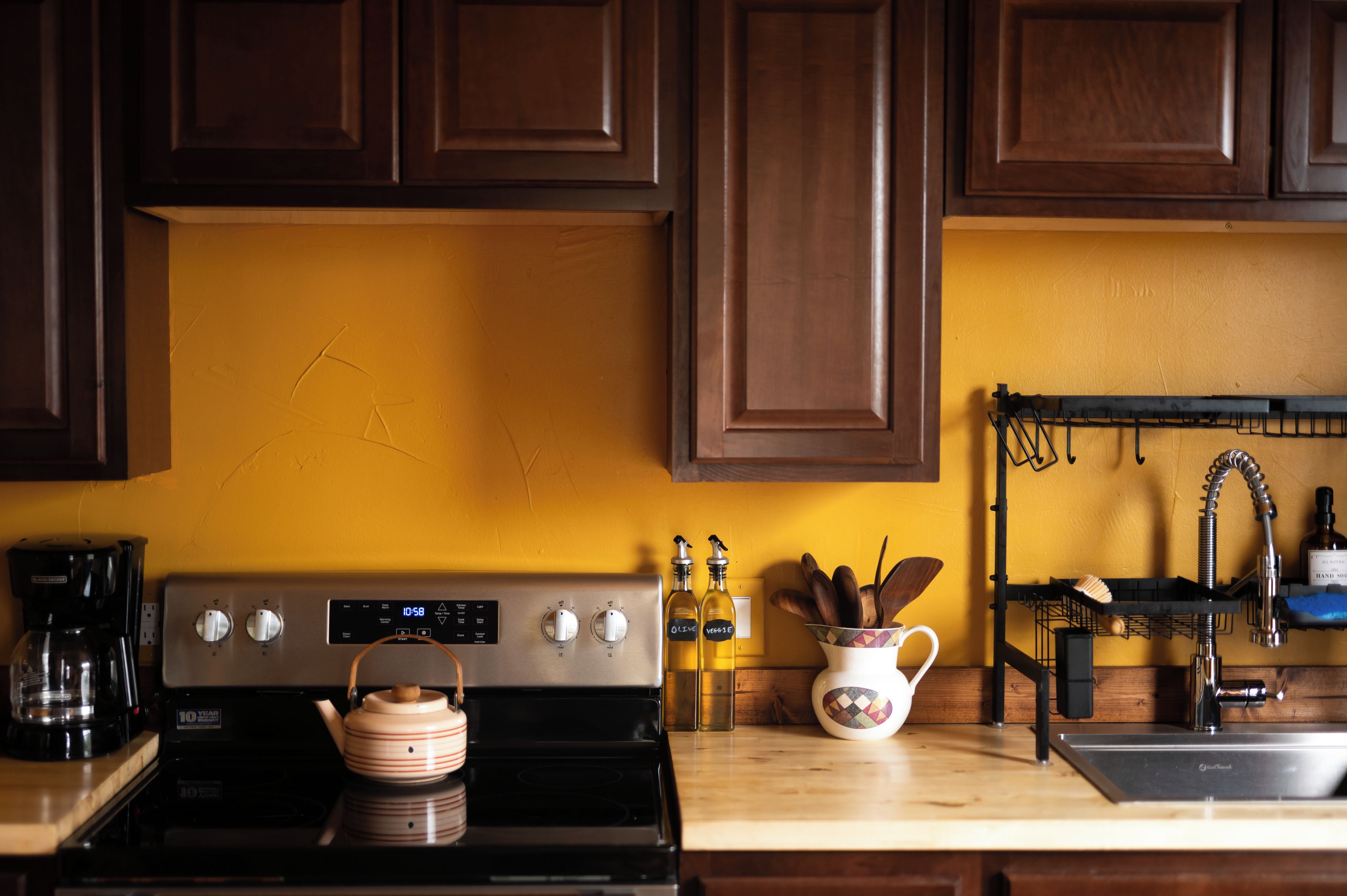A kitchen with a stove, coffee maker, and a kettle. There are bottles labeled 'OIL' and 'VEGGIE' on the counter.