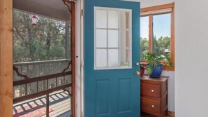 Ein Zimmer mit blauer Tür, Holzboden, Fenster mit Blick auf Bäume und ein kleiner, hölzerner Schminktisch mit einer Vase.