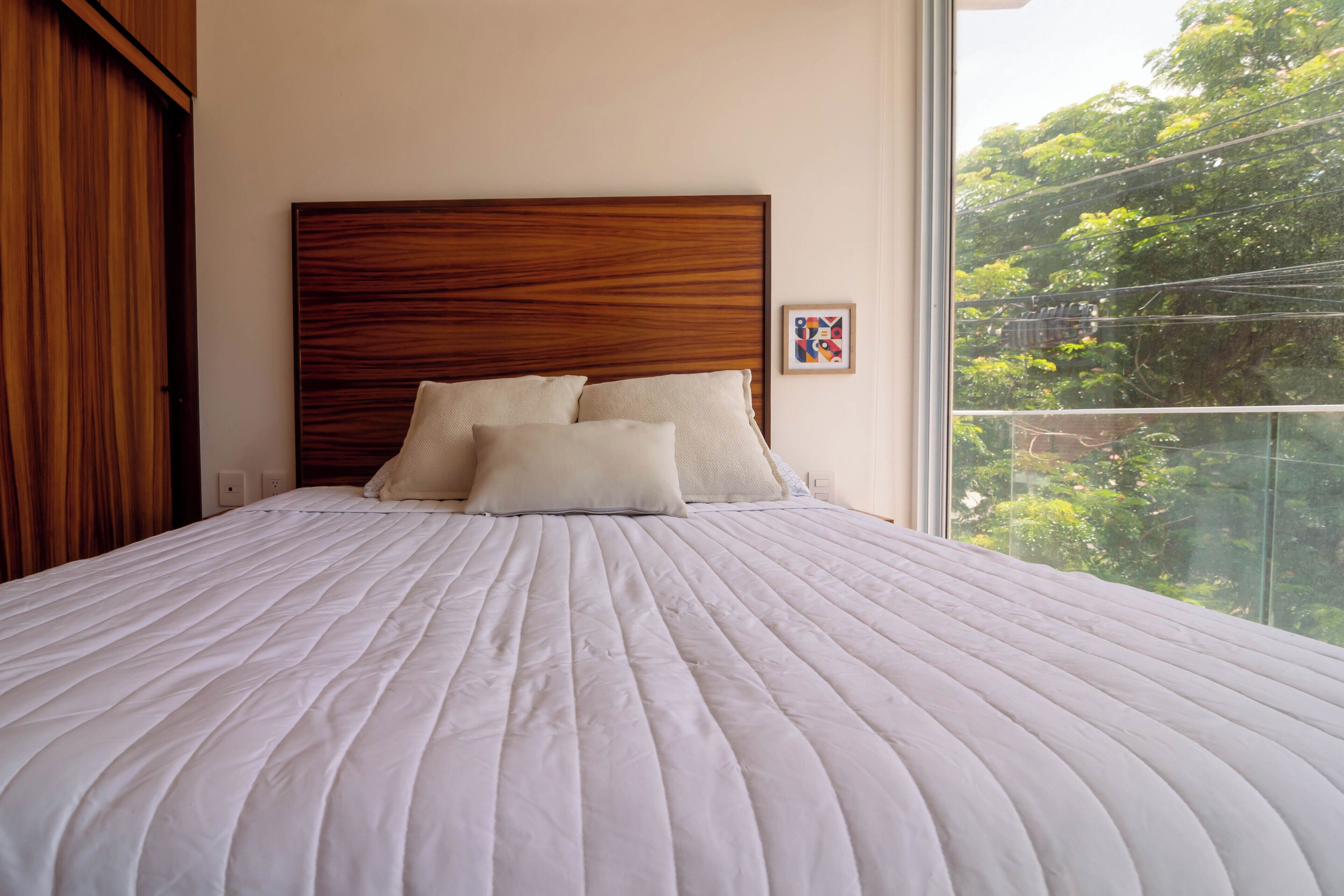 A neatly made bed with a wooden headboard, a window with a view of greenery, and a small framed picture on the wall.