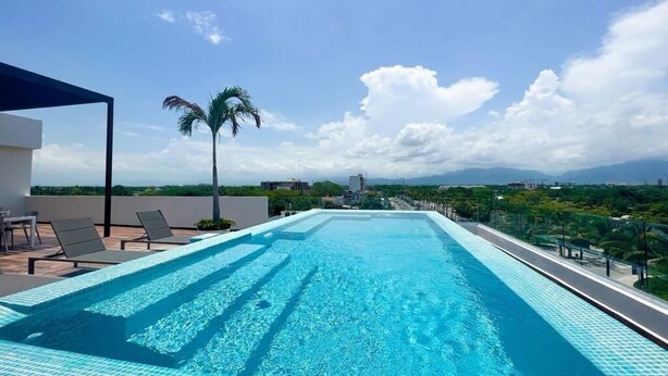 A rooftop infinity pool with clear blue water, surrounded by lounge chairs and palm trees, overlooking a cityscape with mountains in the distance.