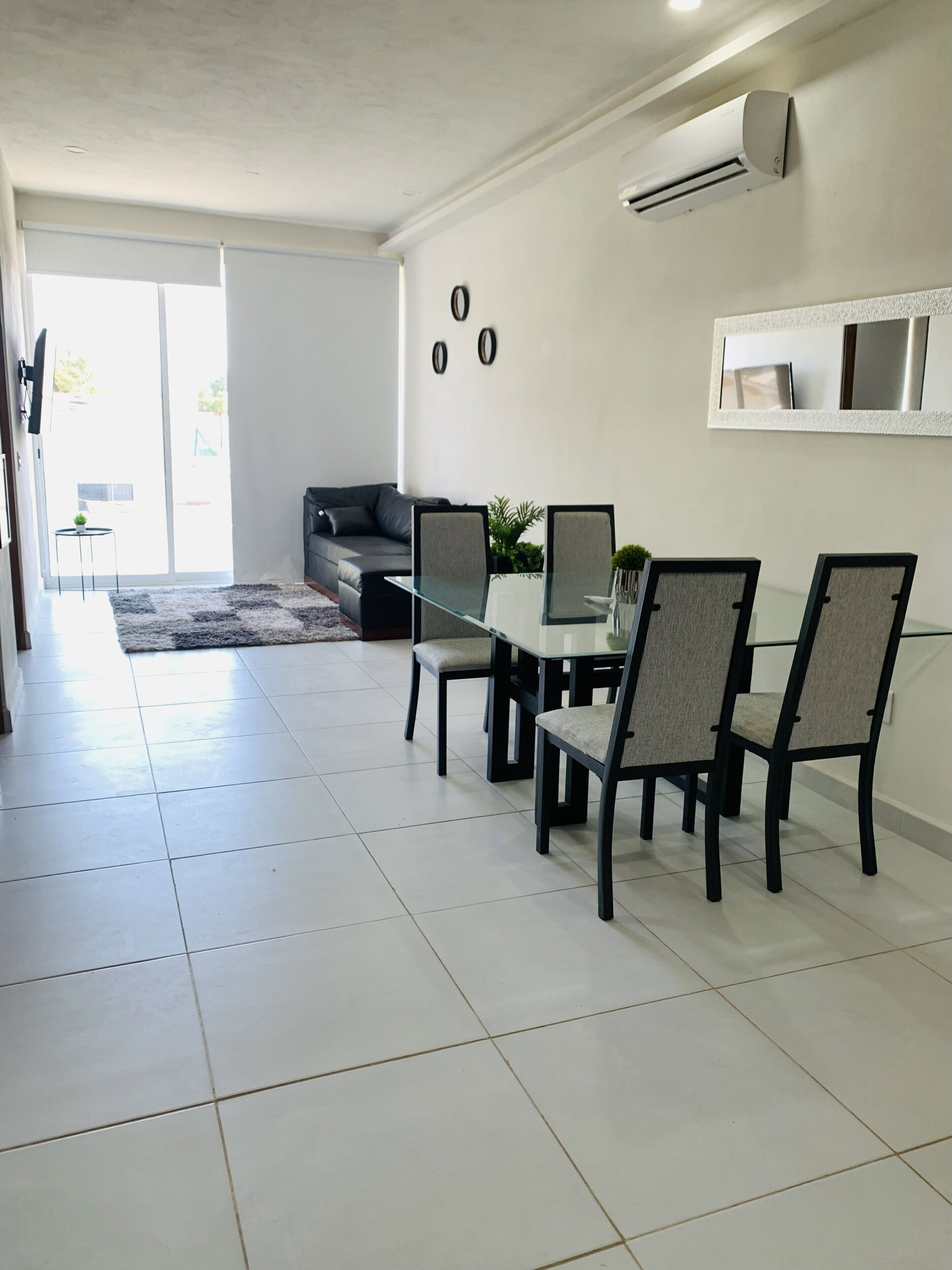 A modern dining area with a glass table and four chairs, a grey sofa, and an air conditioning unit on the wall.