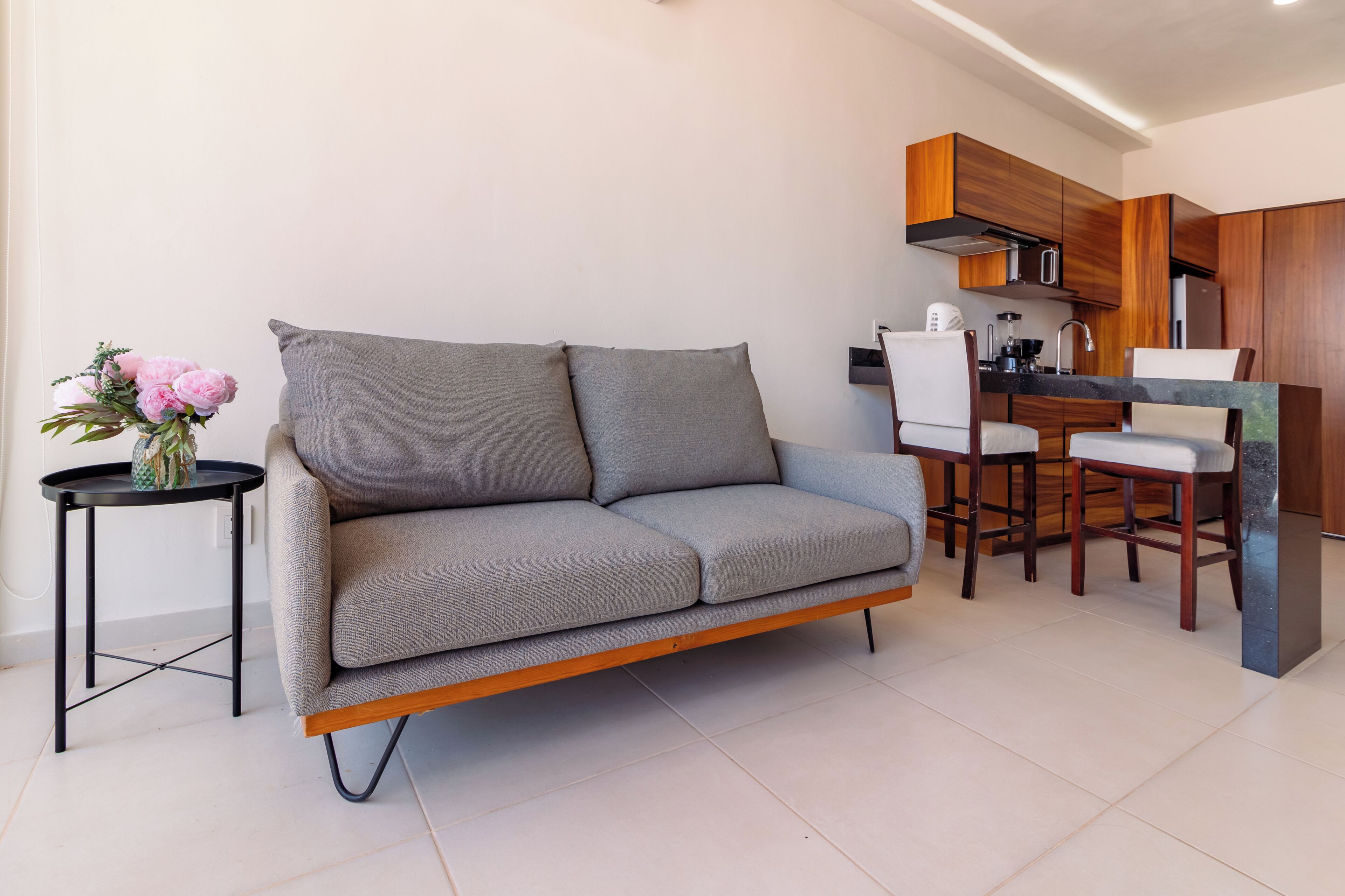A modern living room with a grey sofa, a small round table with a vase, and a kitchen area with wooden cabinets and a dining set.