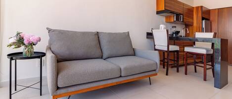 Una sala de estar moderna con un sofá gris, una mesita redonda con un florero, y una zona de cocina con armarios de madera y un juego de comedor.