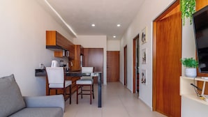A modern kitchen with wooden cabinets, a dining area with a grey sofa, and a TV on a shelf.