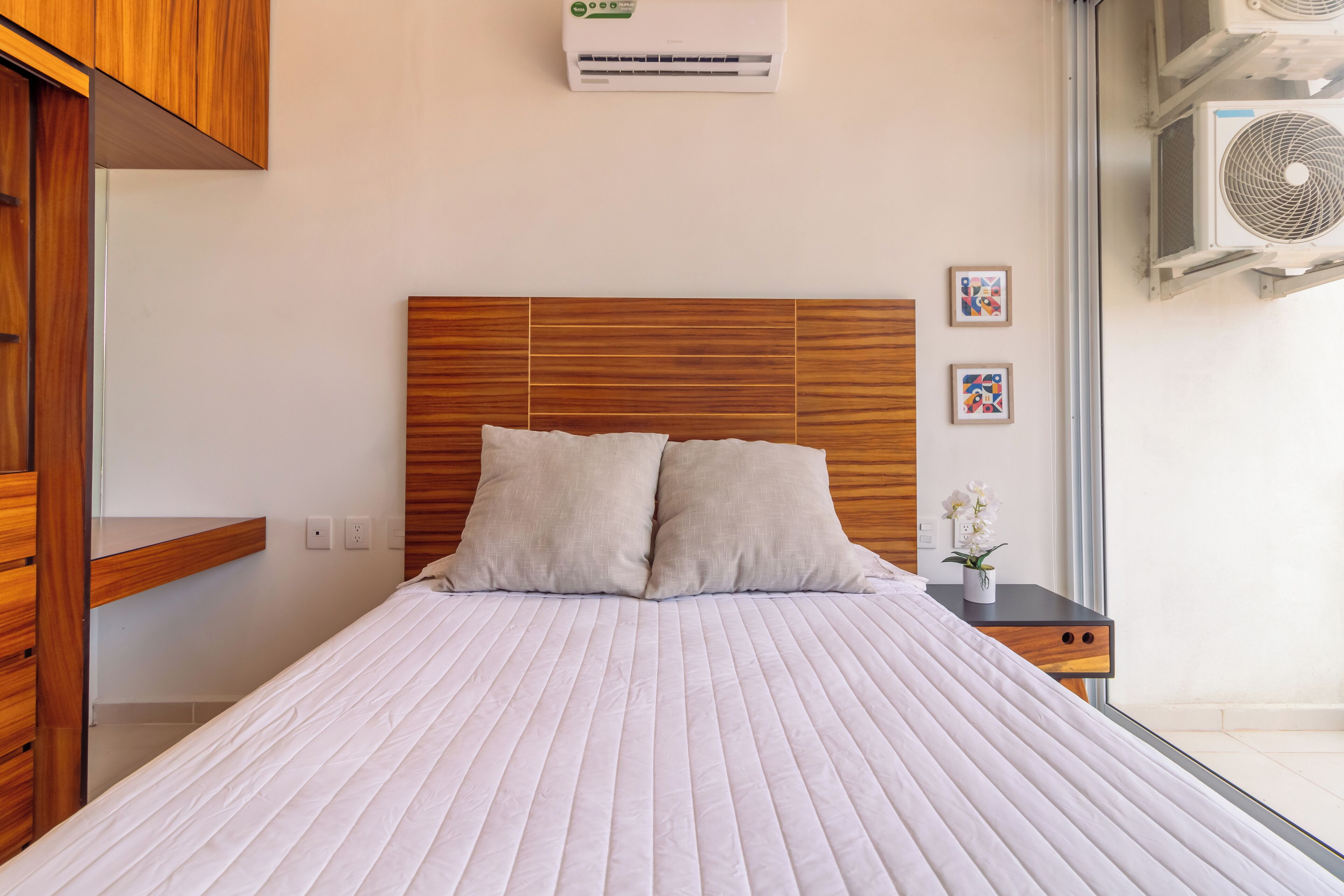 Une chambre à coucher avec une tête de lit en bois, un couvre-lit blanc, deux oreillers, une table de chevet ornée d’un vase de fleurs et un appareil de climatisation placé au-dessus du lit.