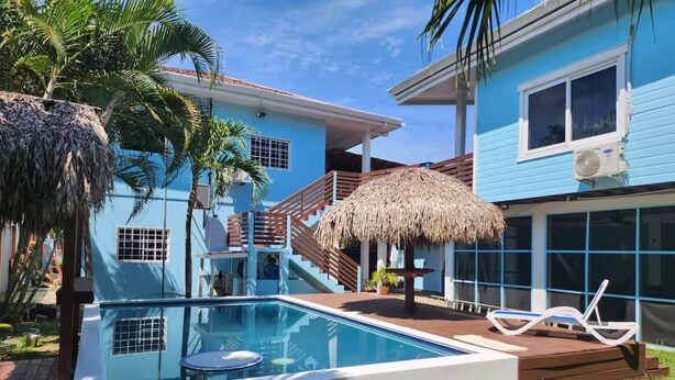 Area piscina con ombrellone di paglia, una poltrona da relax e una scala che conduce a un edificio blu con una finestra e un condizionatore.