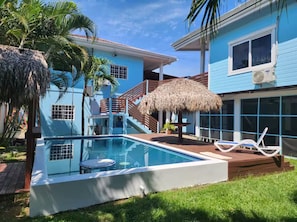 A pool area with a thatched umbrella, a lounge chair, and a staircase leading to a blue building with a window and an air conditioning unit.