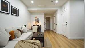 A modern living room with a sofa, coffee table, and a dining area with a table and chairs. There are white walls, wooden flooring, and a door leading to another room. - CASA BOHO I Cozy Boho Retreat in NW Calgary (Calgary)