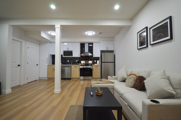Una cocina moderna con armarios blancos, una encimera negra y electrodomésticos de acero inoxidable. En la sala de estar, hay un sofá blanco con cojines y una mesa de centro negra.
