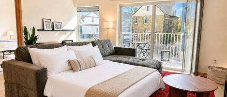 A bedroom with a bed, two sofas, a wooden table, a red rug, and a balcony with a view of houses.