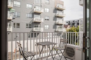 En balkong med ett bord och stolar, en luftkonditioneringsanläggning från Mitsubishi Electric och utsikt över en grå byggnad med flera balkonger.