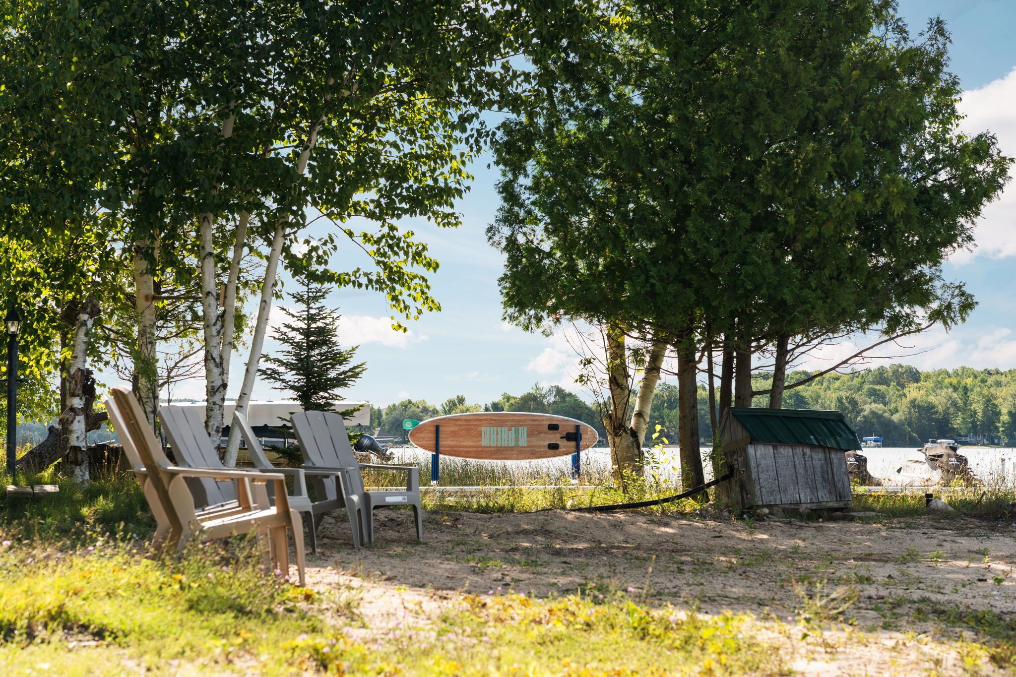 Un paisaje junto al lago con sillas de madera, una tabla de surf de madera y un cobertizo pequeño.