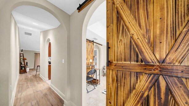 A hallway with wooden flooring and white walls, leading to a room with a large wooden door. - Modern Getaway-Private Tub & Game Room (Ocala)