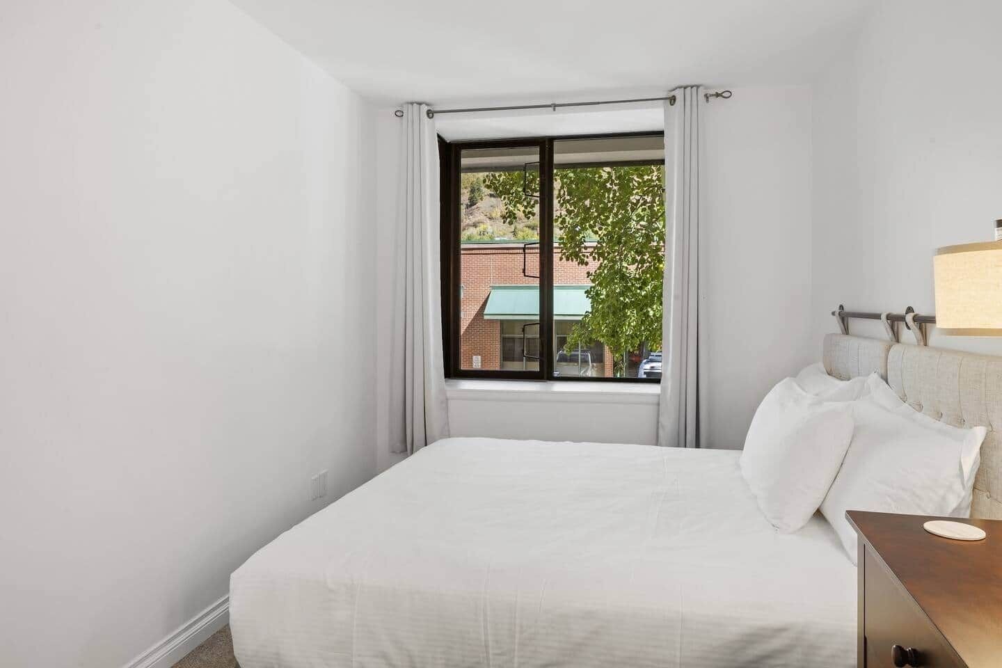 Une chambre d’hôtel avec un grand lit, une table de chevet, une lampe et une fenêtre donnant sur des immeubles et des arbres.