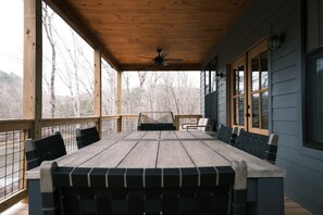 Un espace extérieur abrité, avec une table et des chaises en bois, de grandes fenêtres et une vue sur les arbres.