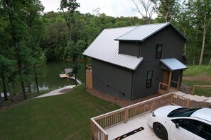A two-story cabin with a metal roof, a deck, and a dock extending into a lake, surrounded by trees.
