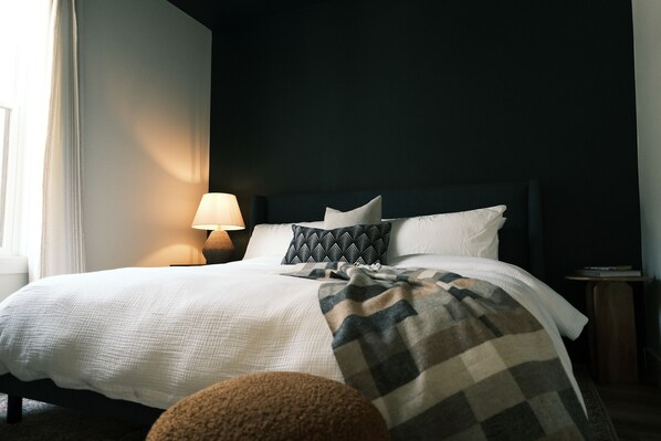 Un lit bien fait, avec une couette blanche et un plaid à carreaux, une lampe de chevet et une petite table de nuit en bois sur laquelle repose un livre.