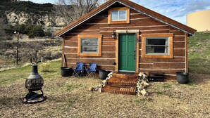 Eine Holzhütte mit grüner Tür, zwei Fenstern und einer kleinen Veranda mit Stühlen.