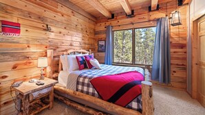 Un dormitori de cabana de fusta, amb un llit, una tauleta de nit, una làmpada i una finestra amb cortines.