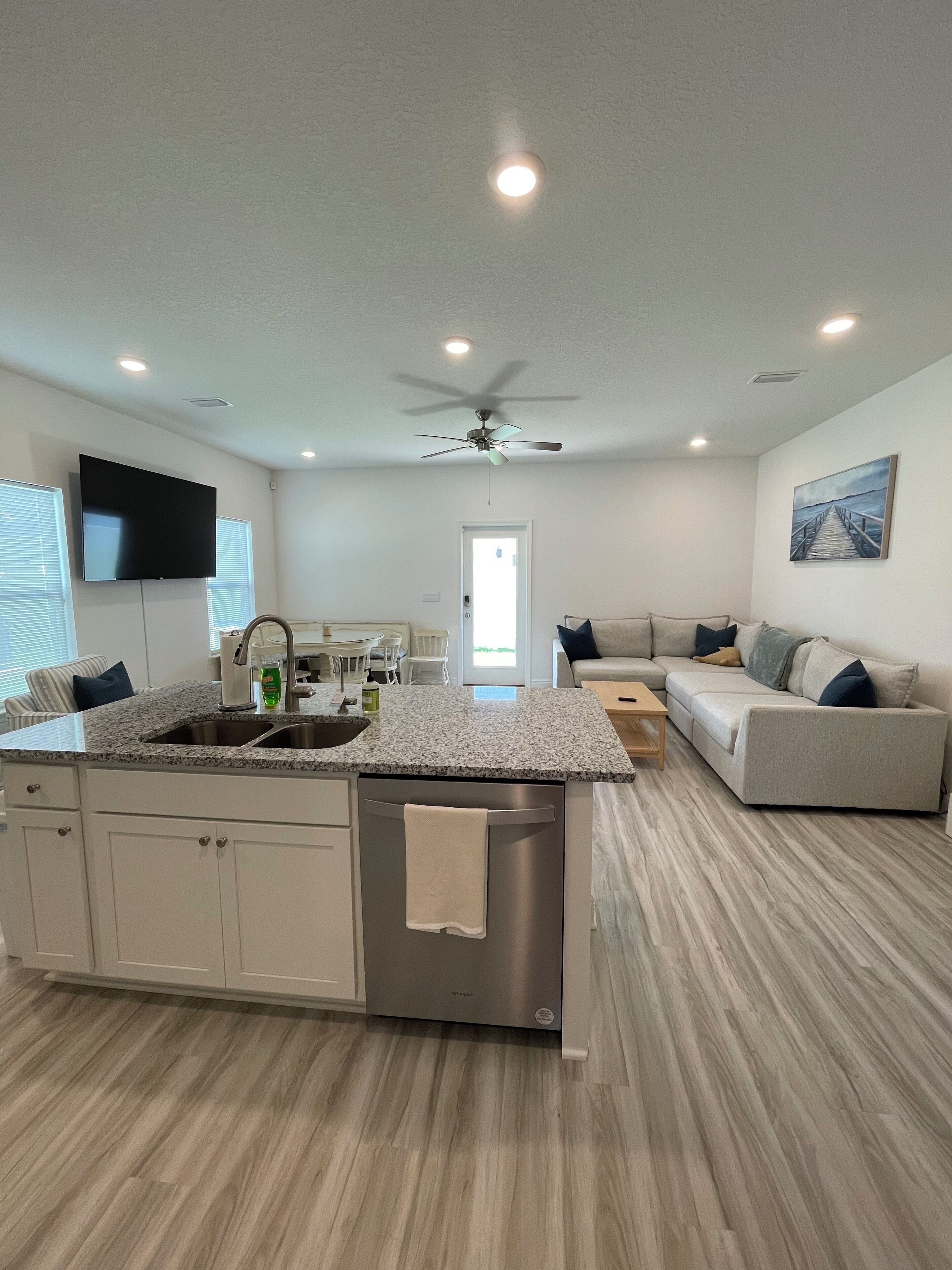 A modern kitchen with a central island, a dining area with a table and chairs, a living room with a sofa, and a ceiling fan.