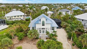 A large, single-story house with a blue roof and white walls, surrounded by lush greenery and palm trees. - Stunning pool home steps to beach! REDUCED! (Sanibel)