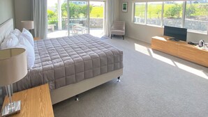 A spacious bedroom with a large bed, a TV on a wooden console, and a balcony with a view of greenery.