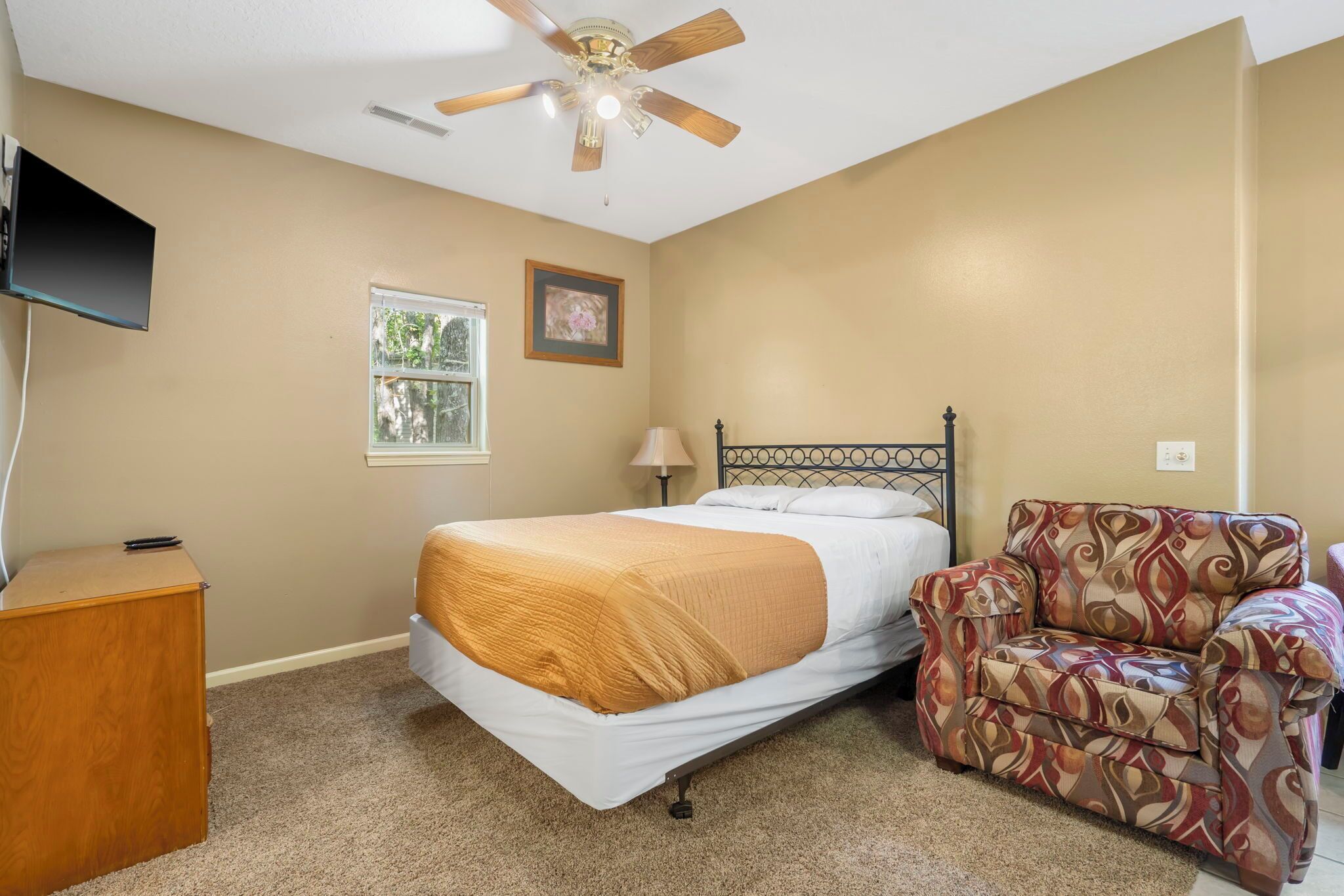 A bedroom with a bed, a chair, a desk, a ceiling fan, a TV, a window, and a framed picture on the wall.