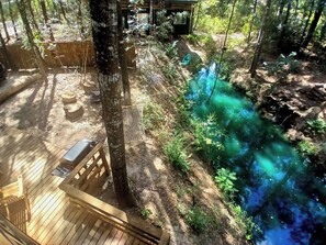 Une terrasse en bois avec un banc, un arbre et un ruisseau aux eaux bleues.
