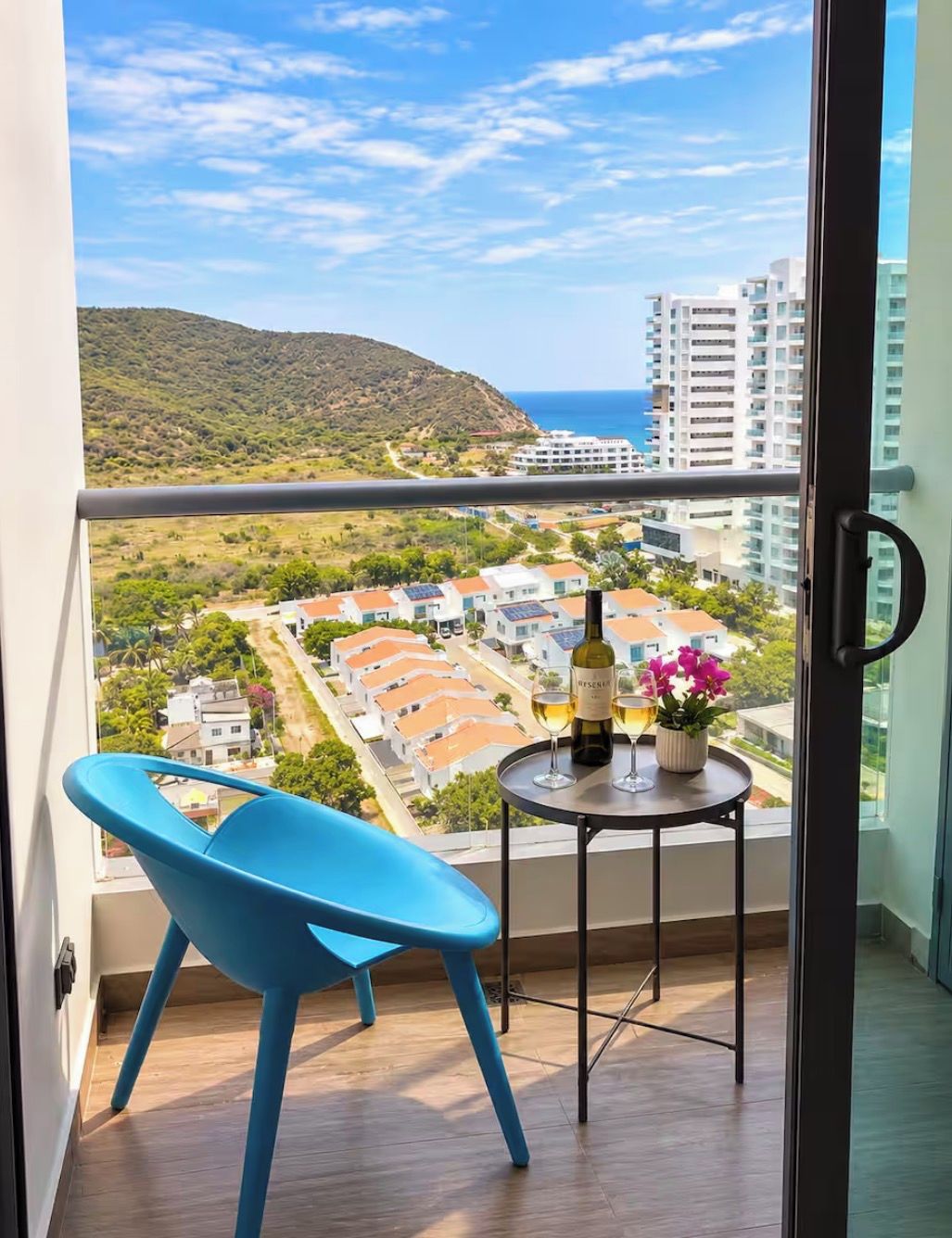 A balcony with a view of a coastal town, a table with a bottle and glasses, and a blue chair.