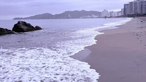 A beach with waves, a rocky outcrop, and a row of buildings in the background. - Cozy Apartment One Block from the Ocean in Salguero Beach, Santa Marta (Gaira)