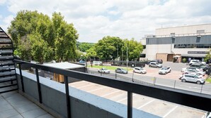 Une vue depuis un balcon donnant sur un parking où se trouvent plusieurs voitures, un café et, au loin, des arbres.