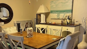 A dining area with a wooden table, white chairs, a ceiling fan, and a painting of a beach scene. - Valerie’s Villa - free WiFi - 3 tv’s - 15 mins to beach - 30 second walk to pool (Fort Myers)