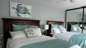 A bedroom with two beds, each with a wooden headboard, white bedding, and teal pillows. There is a wall-mounted artwork of a turtle, a ceiling fan, and a large mirror. - Valerie’s Villa - free WiFi - 3 tv’s - 15 mins to beach - 30 second walk to pool (Fort Myers)