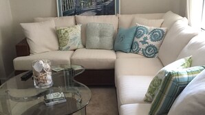 A living room with a beige sectional sofa, a glass coffee table, and a wicker side table. - Valerie’s Villa - free WiFi - 3 tv’s - 15 mins to beach - 30 second walk to pool (Fort Myers)