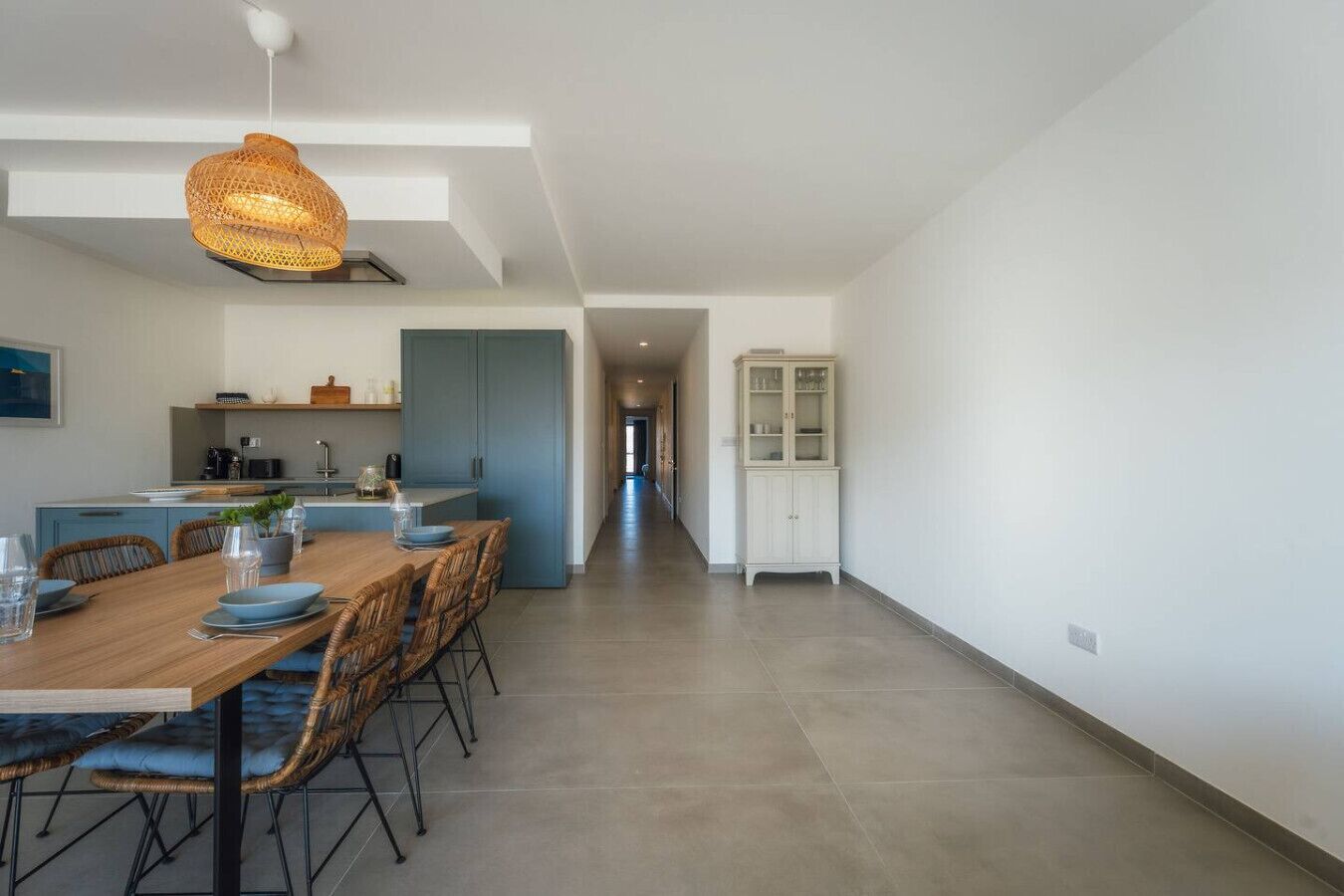 A modern kitchen with a dining area, featuring a wooden table, wicker chairs, and a hanging light fixture.