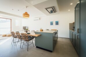 A modern kitchen with a central island, dining area with wicker chairs, and a large window providing natural light. - Qala Skyview Penthouse Pool Sunsets BBQ Area (Qala)